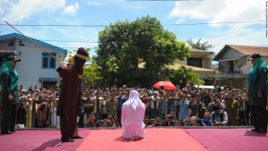 The man in the mask is known as a "religious officer" in the country of Indonesia. A strict Muslim theocracy, the person in white is about to be publicly caned because he was dating before getting married.